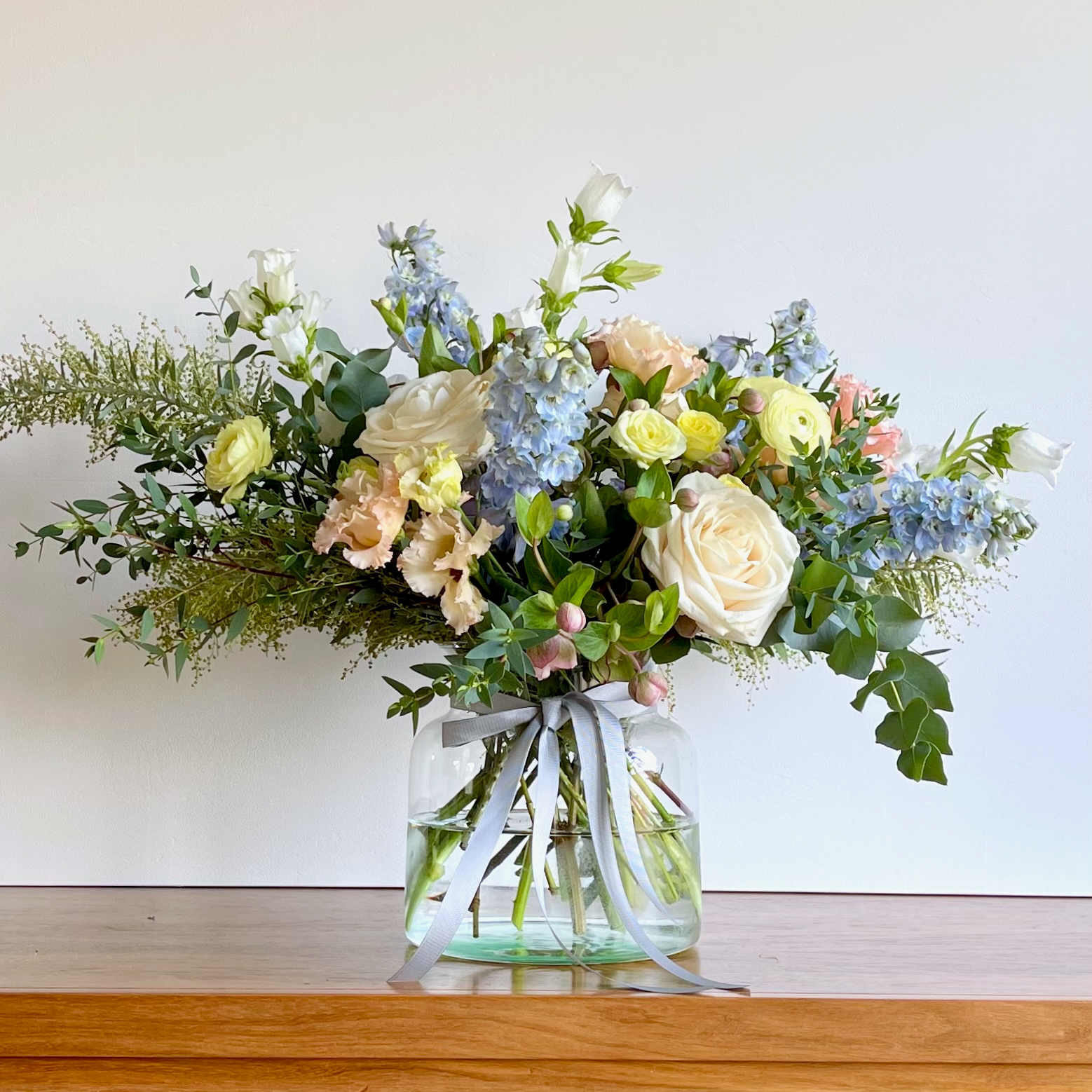 A Mother's Day bouquet with a mix of pale yellow, soft white, pink, and blue flowers, arranged in a clear glass vase, with green foliage.