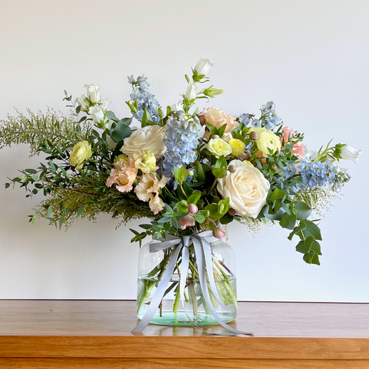 A Mother's Day bouquet with a mix of pale yellow, soft white, pink, and blue flowers, arranged in a clear glass vase, with green foliage.