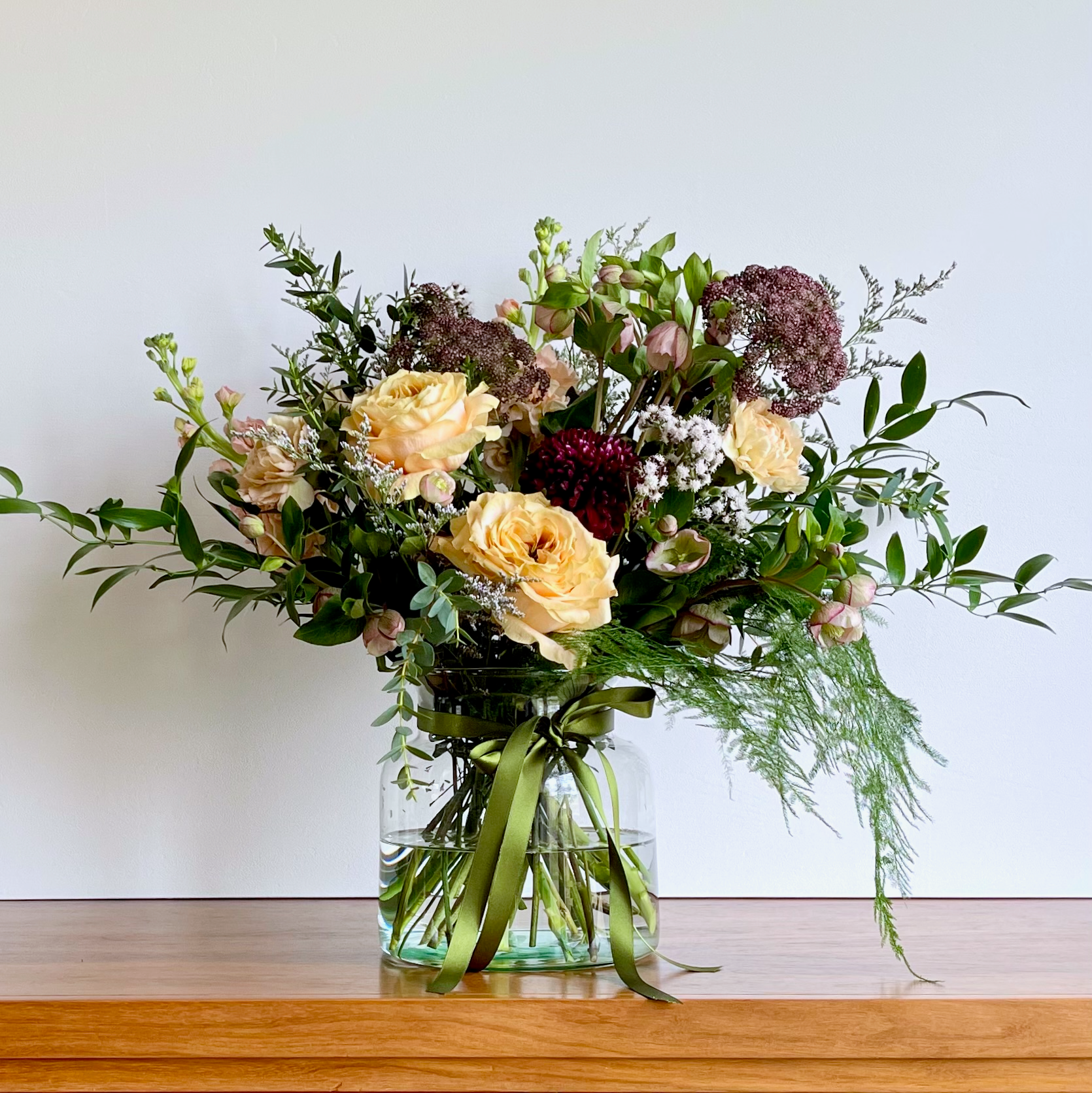 A Mother's Day bouquet with a mix of antique gold, plum, and peachy pink flowers, including eucalyptus and foliage, arranged in a clear glass vase.