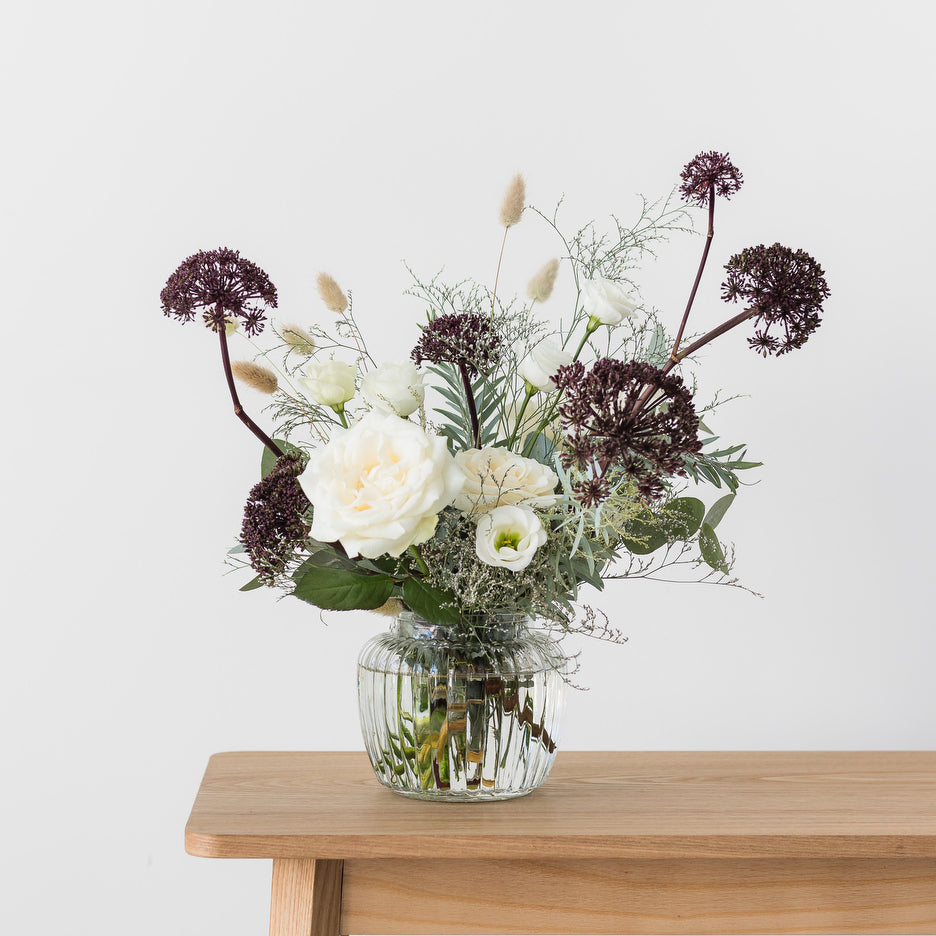A seasonal posy jar arrangement featuring a variety of flowers and foliage in a small ribbed glass vase, placed on a wooden table.